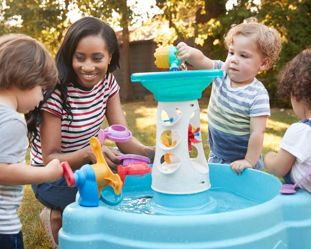 Nursery worker with three toddlers, playing with some toys, in water, outside.