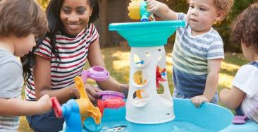 Nursery worker with three toddlers, playing with some toys, in water, outside.